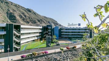Un hotel moderno con varios pisos, zonas recreativas al aire libre y un paisaje de montañas al fondo.