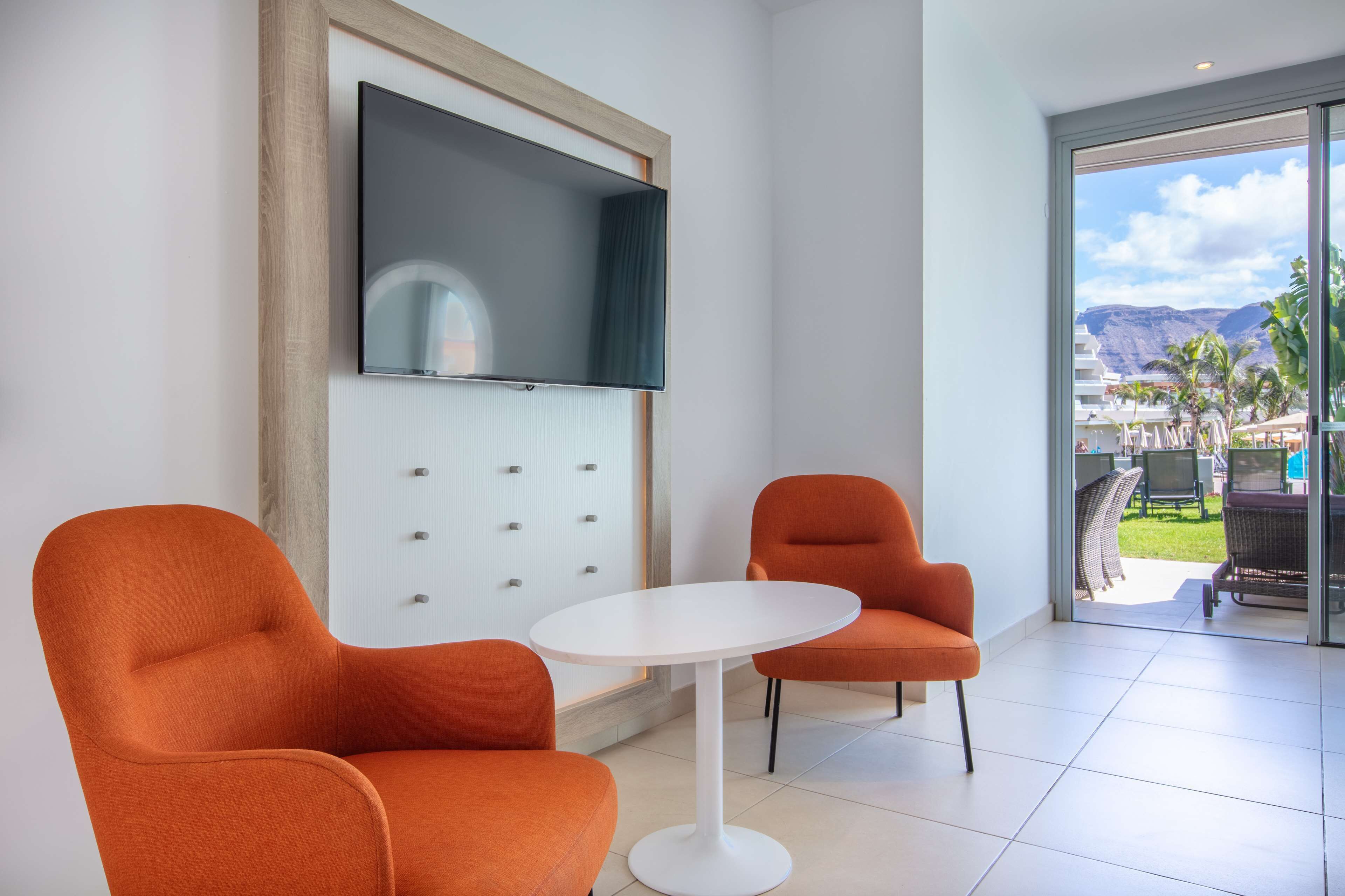 A modern room with a white table and two orange chairs, a flat-screen TV mounted on the wall, and a view of the outdoors through a glass door.