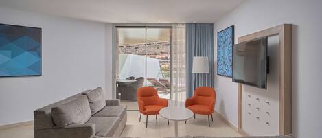 A modern living room with a grey sofa, two orange chairs, a small white table, a TV, and a large window with a view of a building and a mountain.