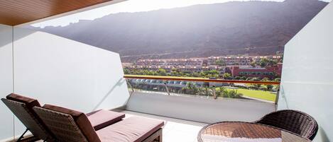 Un balcon agrémenté de meubles en osier, d’une table en verre et offrant une vue sur le paysage urbain et les montagnes.
