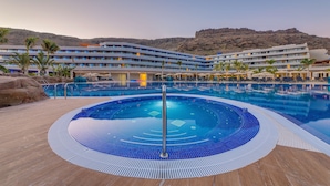 Un hotel con una piscina grande, palmeras y montañas al fondo.