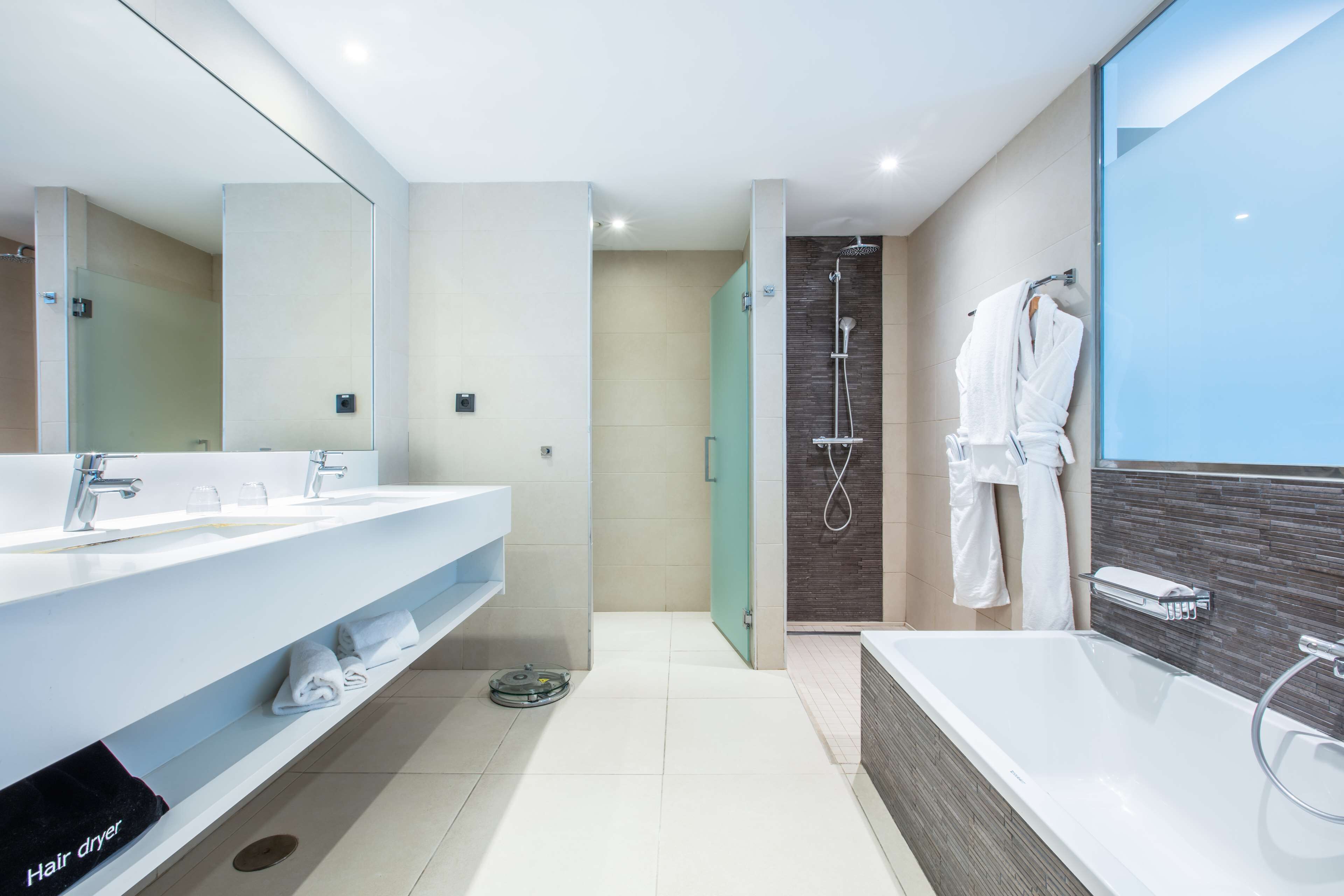 Une salle de bain moderne avec deux lavabos, une grande baignoire, un espace douche et un sèche-cheveux posé sur le sol.