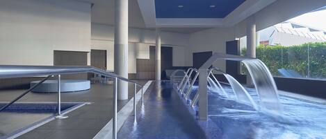 A modern indoor pool area with a long, narrow pool, multiple water jets, and a view of greenery outside.