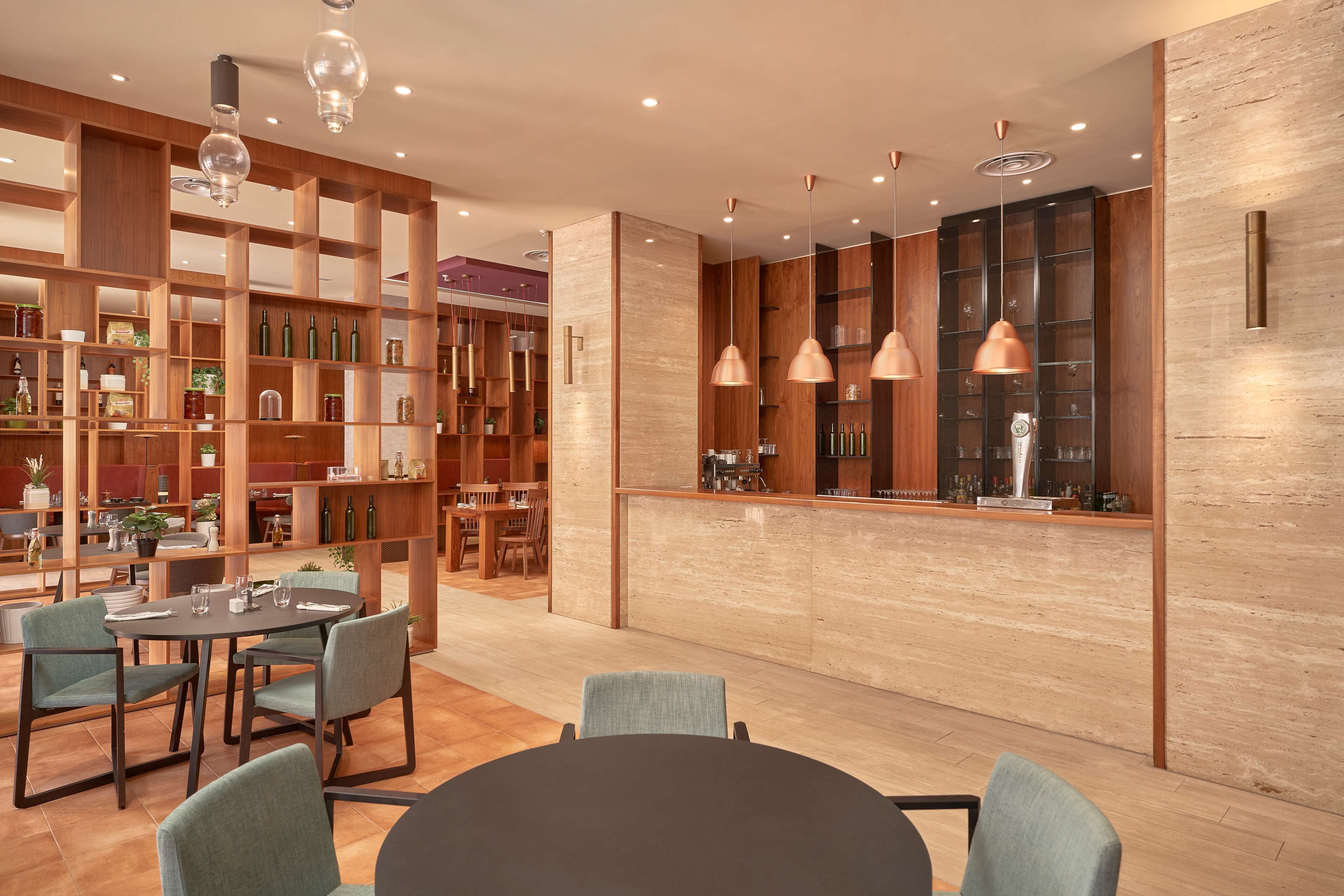 Un interior moderno con un mostrador de recepción de madera, mesas redondas y sillas. Se observan estantes con botellas y adornos.