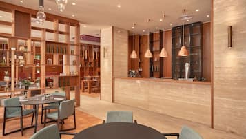 Un interior moderno con un mostrador de recepción de madera, mesas redondas y sillas. Se observan estantes con botellas y adornos.
