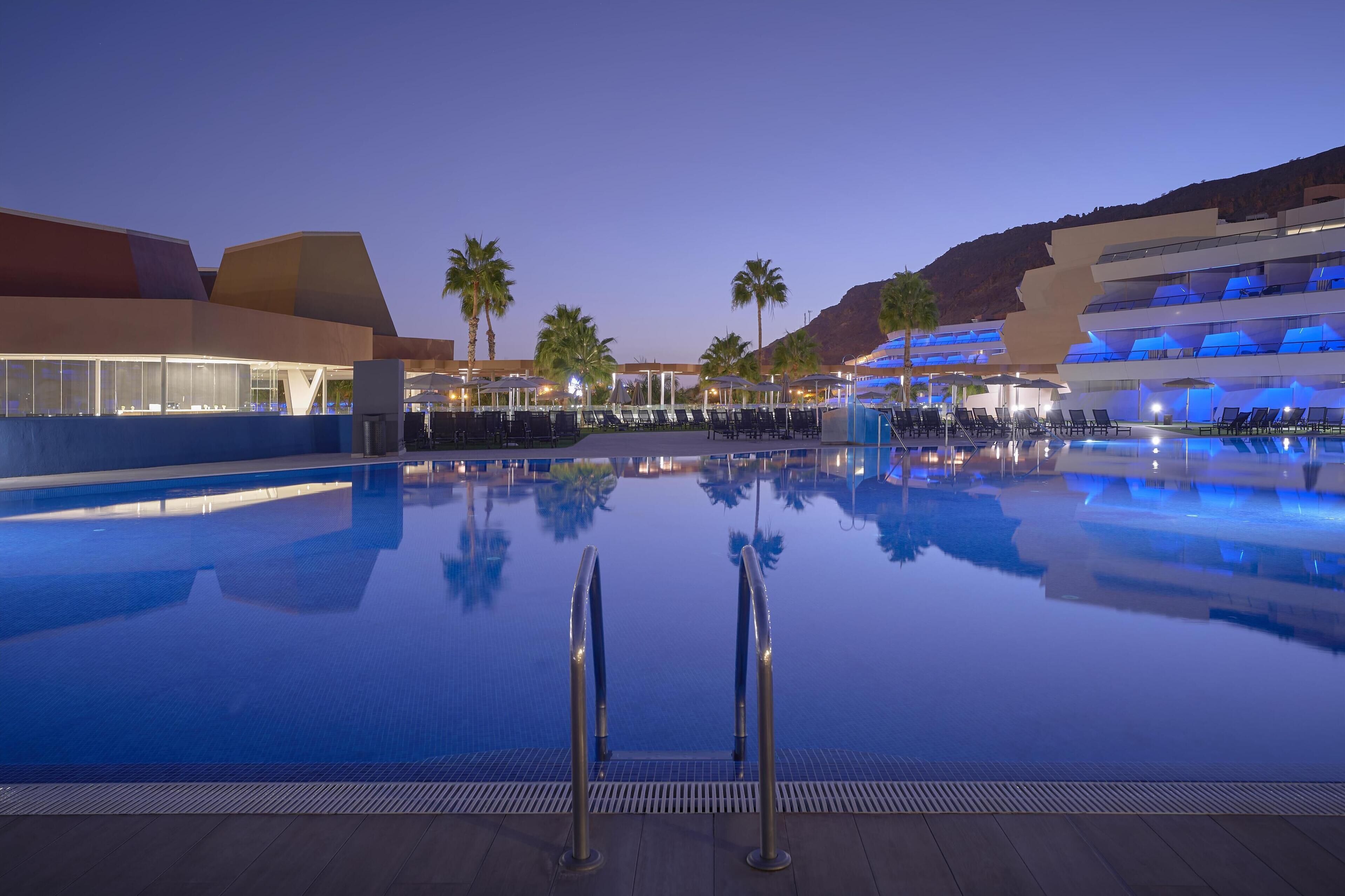 A large outdoor swimming pool with a modern architectural backdrop, palm trees, and a clear sky.