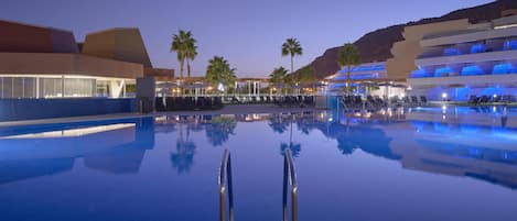 A large outdoor swimming pool with a modern architectural backdrop, palm trees, and a clear sky.