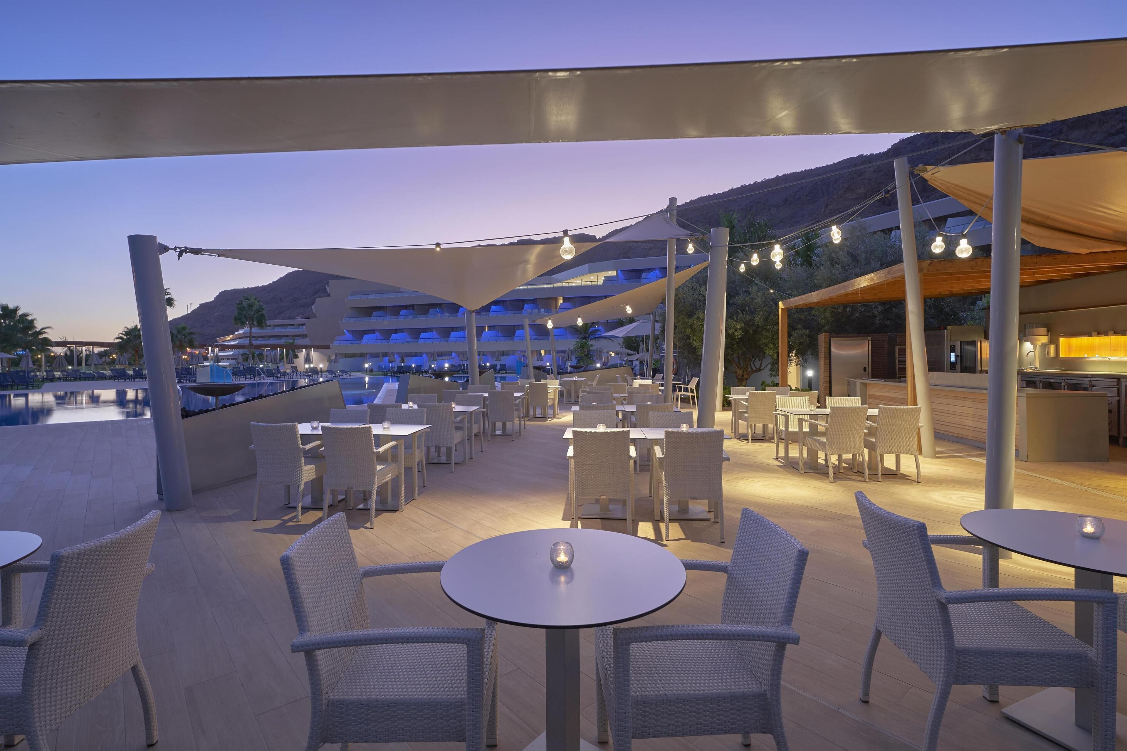 An outdoor dining area with round tables and wicker chairs, shaded by large canopies and string lights.