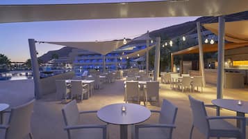 An outdoor dining area with round tables and wicker chairs, shaded by large canopies and string lights.