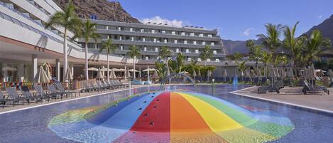 Hotel con piscina que presenta un diseño de arcoíris, palmeras y montañas al fondo.