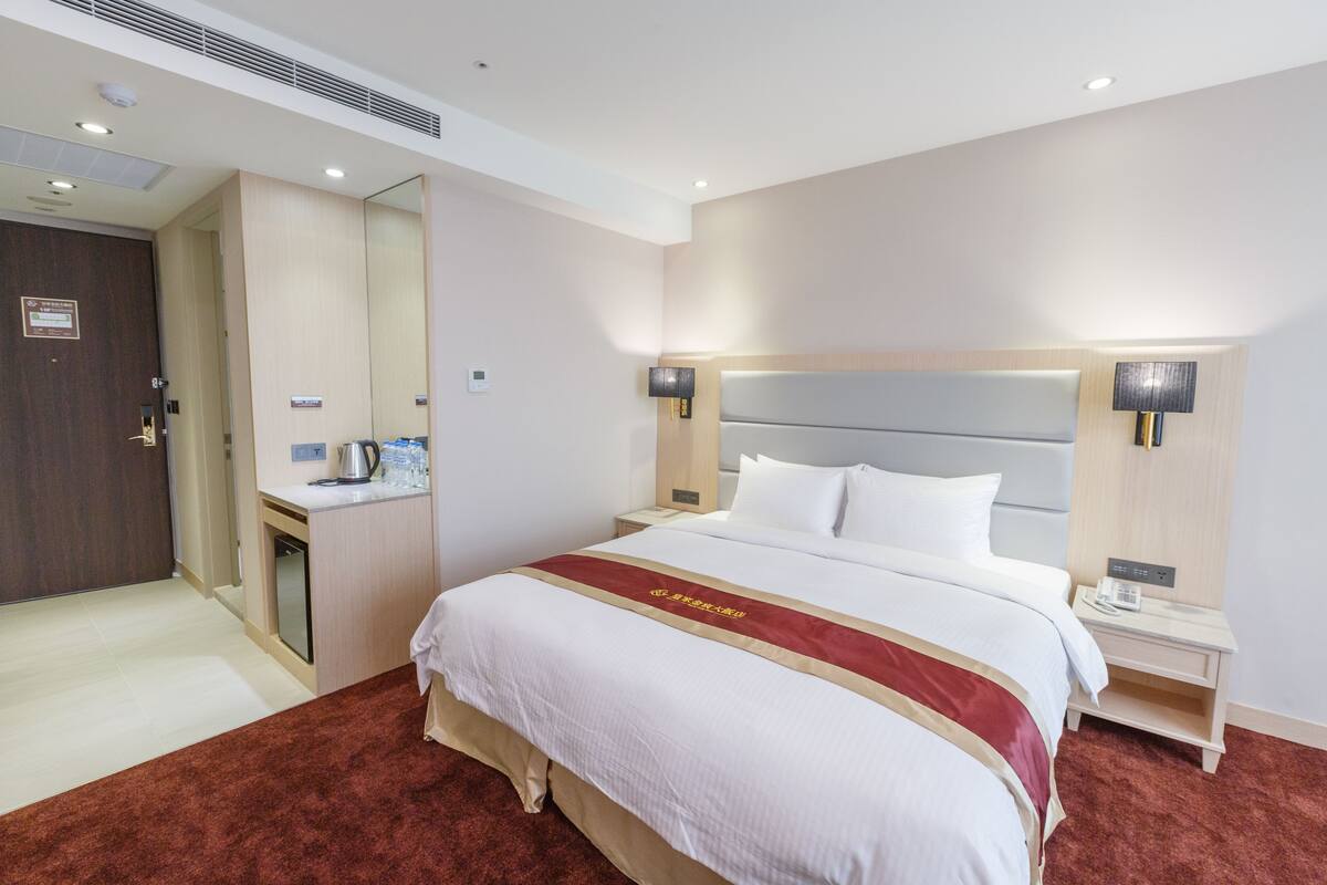 Ein Hotelzimmer mit einem großen Bett, einem Nachttisch mit Lampe, einem kleinen Schreibtisch mit Kaffeemaschine und einem Spiegel.