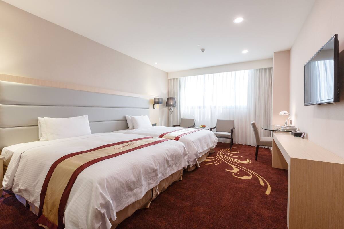 Ein Hotelzimmer mit zwei Betten, einem Schreibtisch, einem Sessel und einem an der Wand befestigten Fernseher.