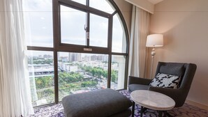 A hotel room with a large window offering a city view, a grey armchair with a floral pillow, a small round table, and a patterned carpet. - Royal Gold Hotel (Kaohsiung)