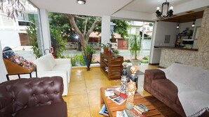 Un vaste espace de vie comprenant un canapé blanc, un fauteuil en cuir marron et un divan marron. On y trouve une table basse en bois avec des magazines et une statue décorative. Une bibliothèque en bois et une plante en pot sont également visibles.