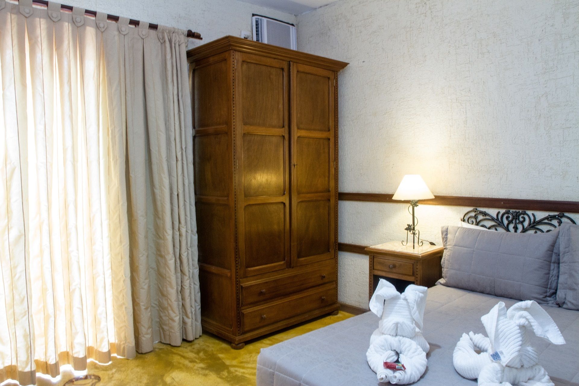 Ein Hotelzimmer mit Holzschrank, einem Bett mit zwei Kissen, einem Nachttisch mit Lampe und einem Vorhangfenster.