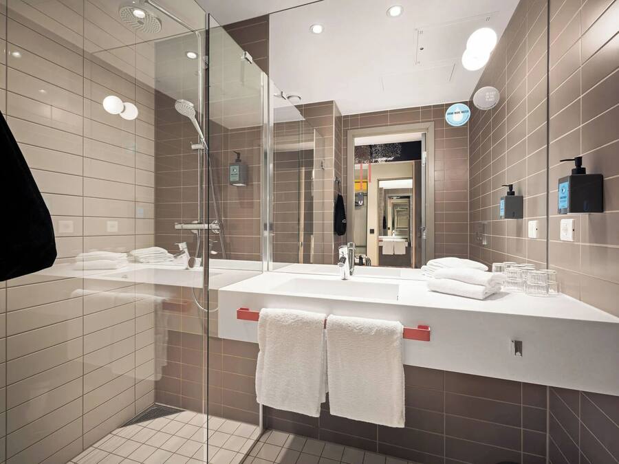 Un baño moderno con cabina de ducha de vidrio, un lavabo blanco con dispensador de jabón rojo y un espejo grande.