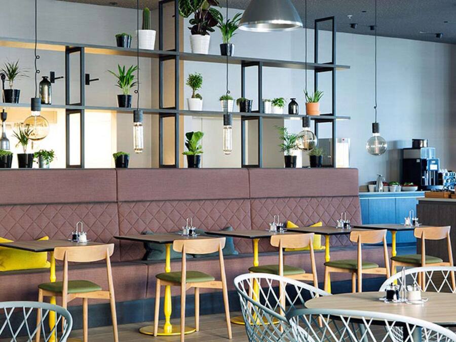 Um restaurante moderno com mesas e cadeiras de madeira, um banco acolchoado e prateleiras com plantas em vasos.