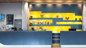 Zona de bar moderna con una decoración azul y amarilla, que incluye una barra, estantes con botellas y una pantalla digital.
