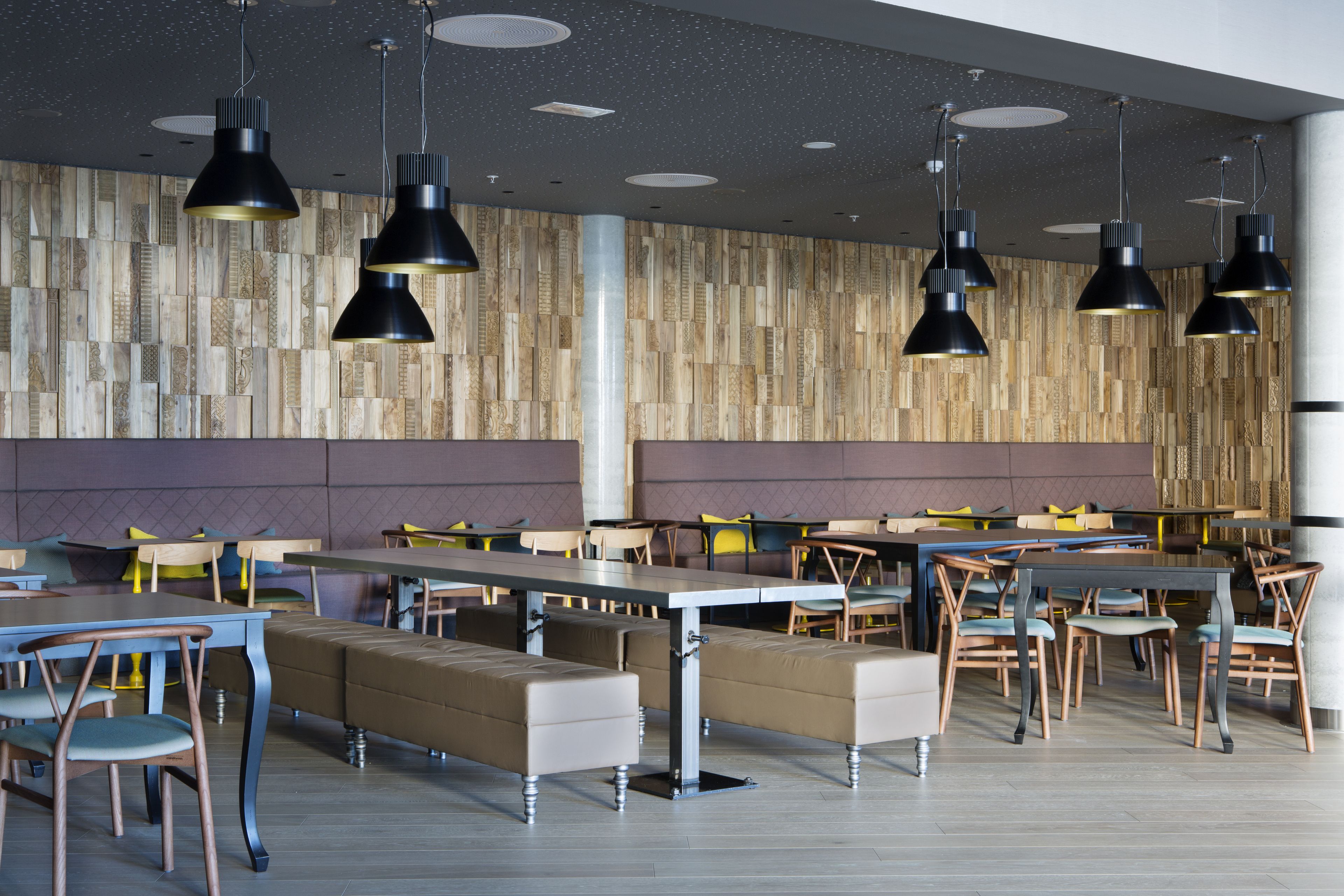 A modern dining area with multiple tables and benches, wooden panel walls, and pendant lighting.
