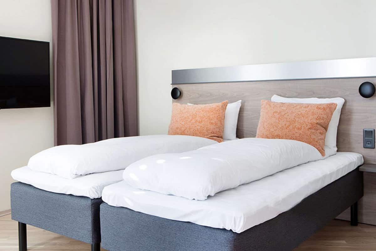 Una cama doble con sábanas blancas y dos almohadas naranjas, un cabecero de madera, un televisor de pantalla plana montado en la pared y una cortina oscura.