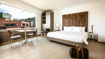 A hotel room with a large bed, a desk with chairs, a small table, a lamp, and a view of buildings outside.