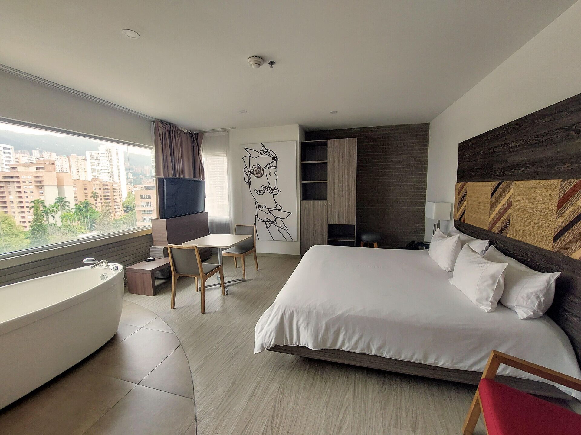 A modern hotel room with a large bed, a bathtub, a dining area, and a city view.