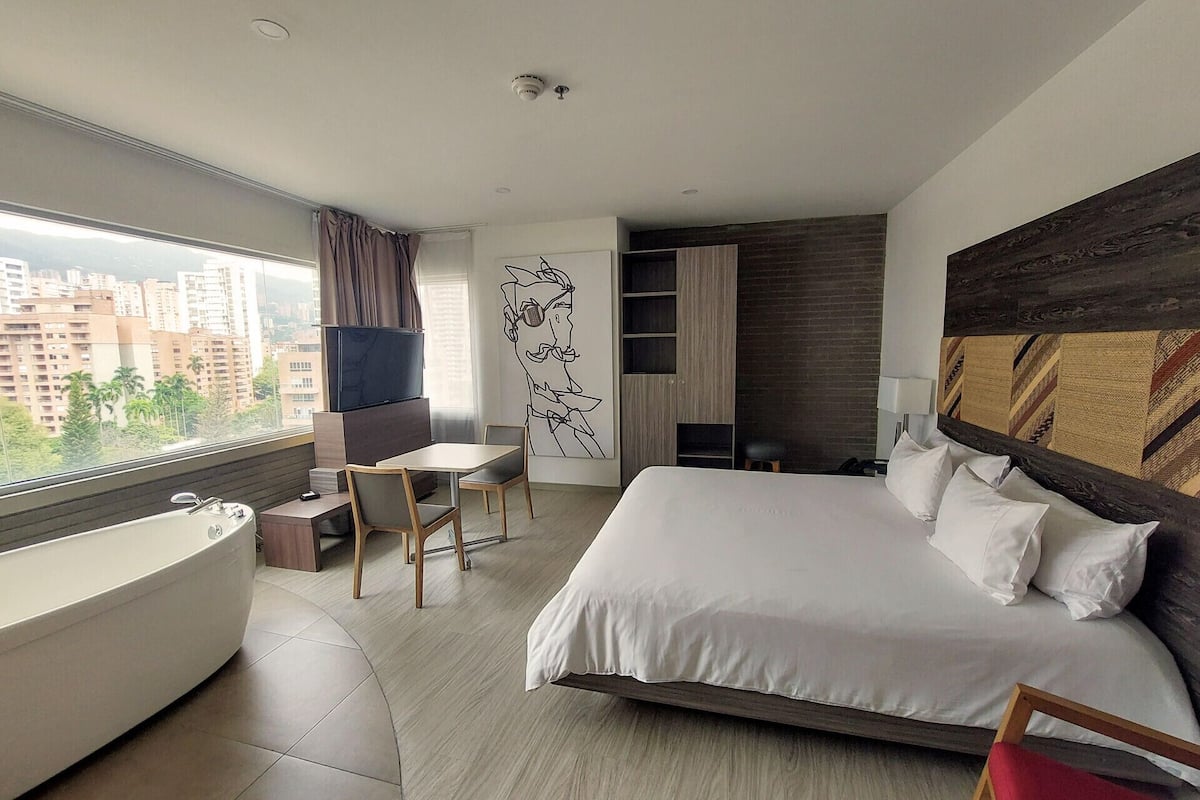 A modern hotel room with a large bed, a bathtub, a dining area, and a city view.