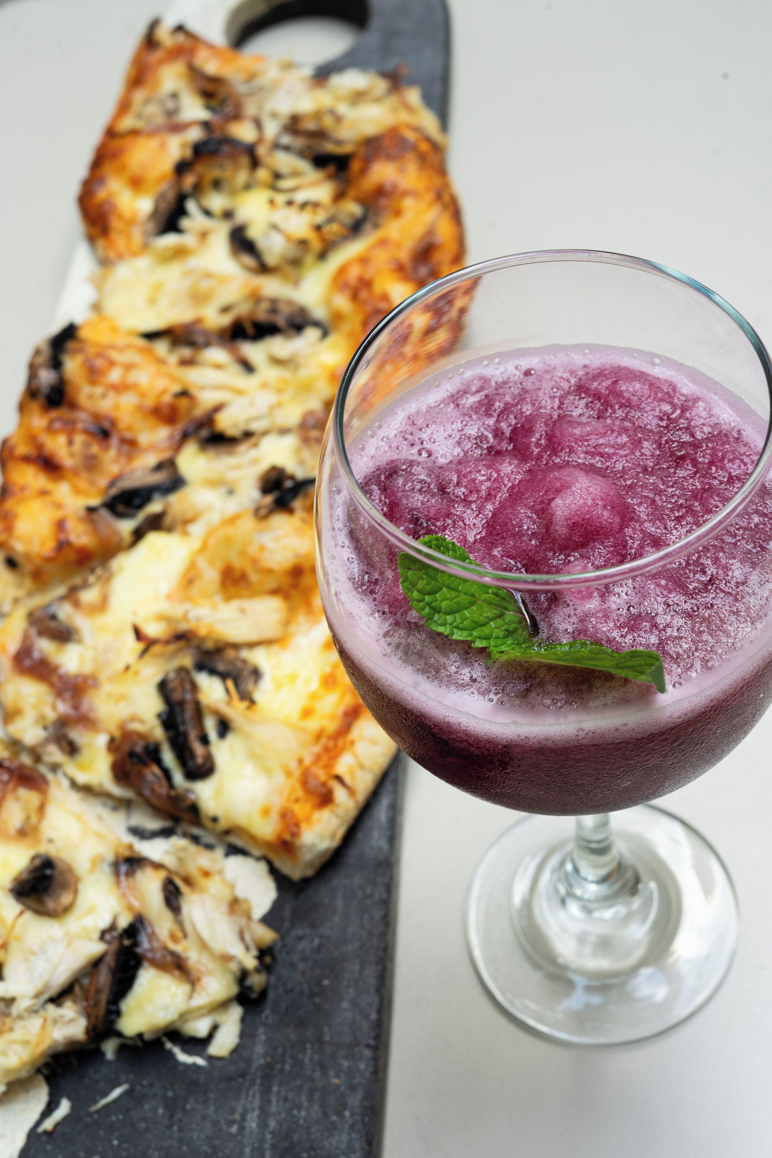 Un verre d’une boisson violette, décoré d’une feuille de menthe, accompagné d’une pizza au fromage et aux champignons.