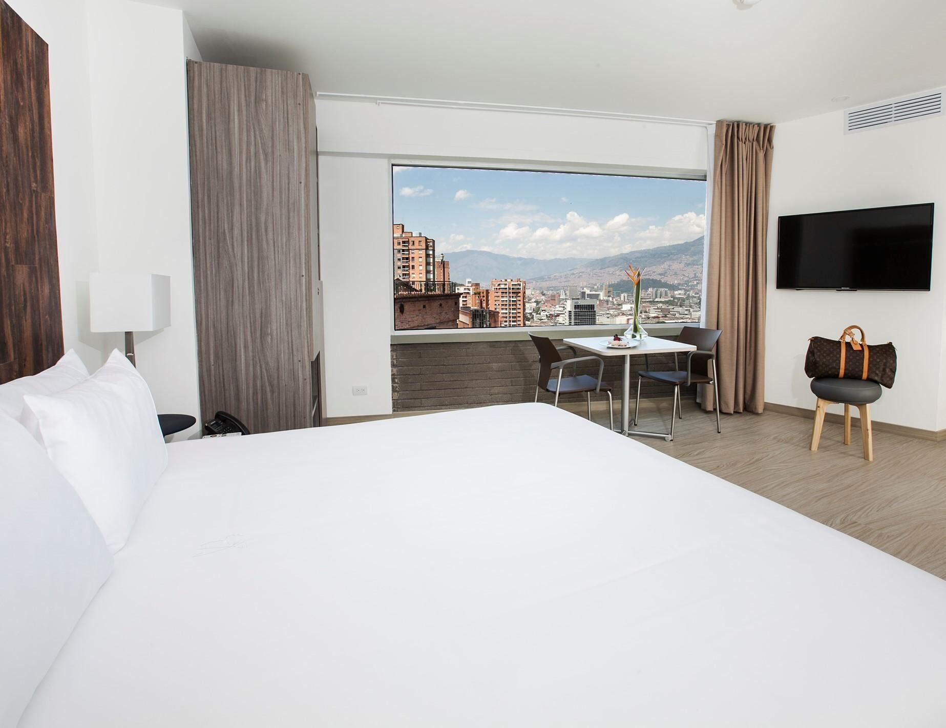 A hotel room with a large bed, a desk with two chairs, a television, and a view of a cityscape.