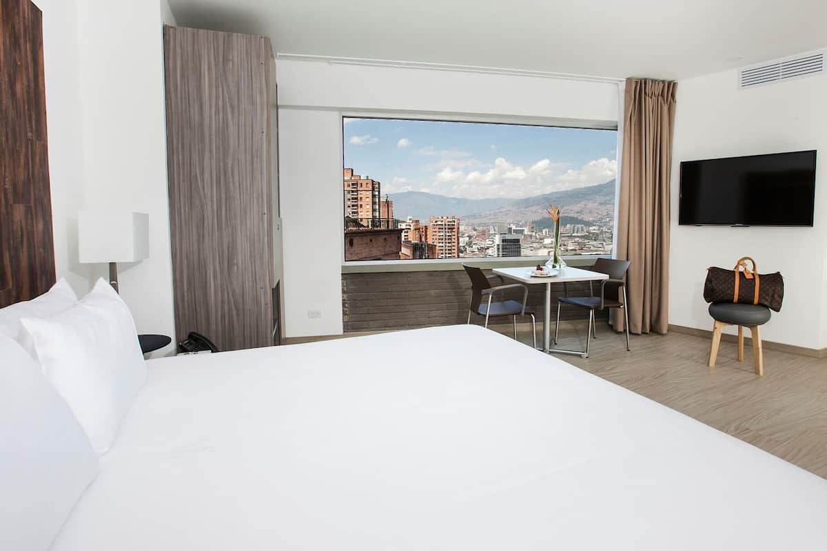 A hotel room with a large bed, a desk with two chairs, a television, and a view of a cityscape.