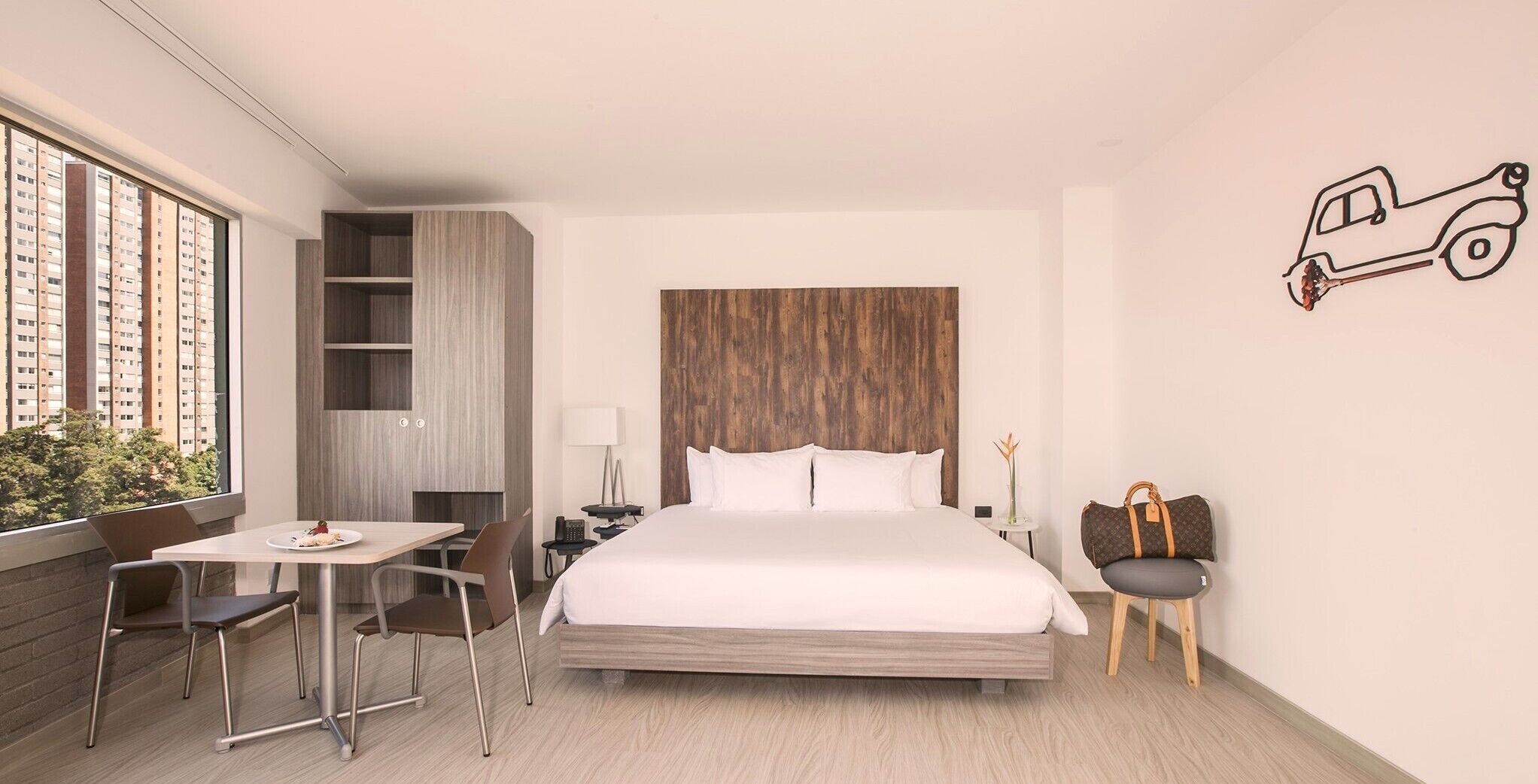 Una habitación de hotel moderna con una cama grande, un escritorio con una silla, una mesita con una lámpara y vistas a edificios altos.