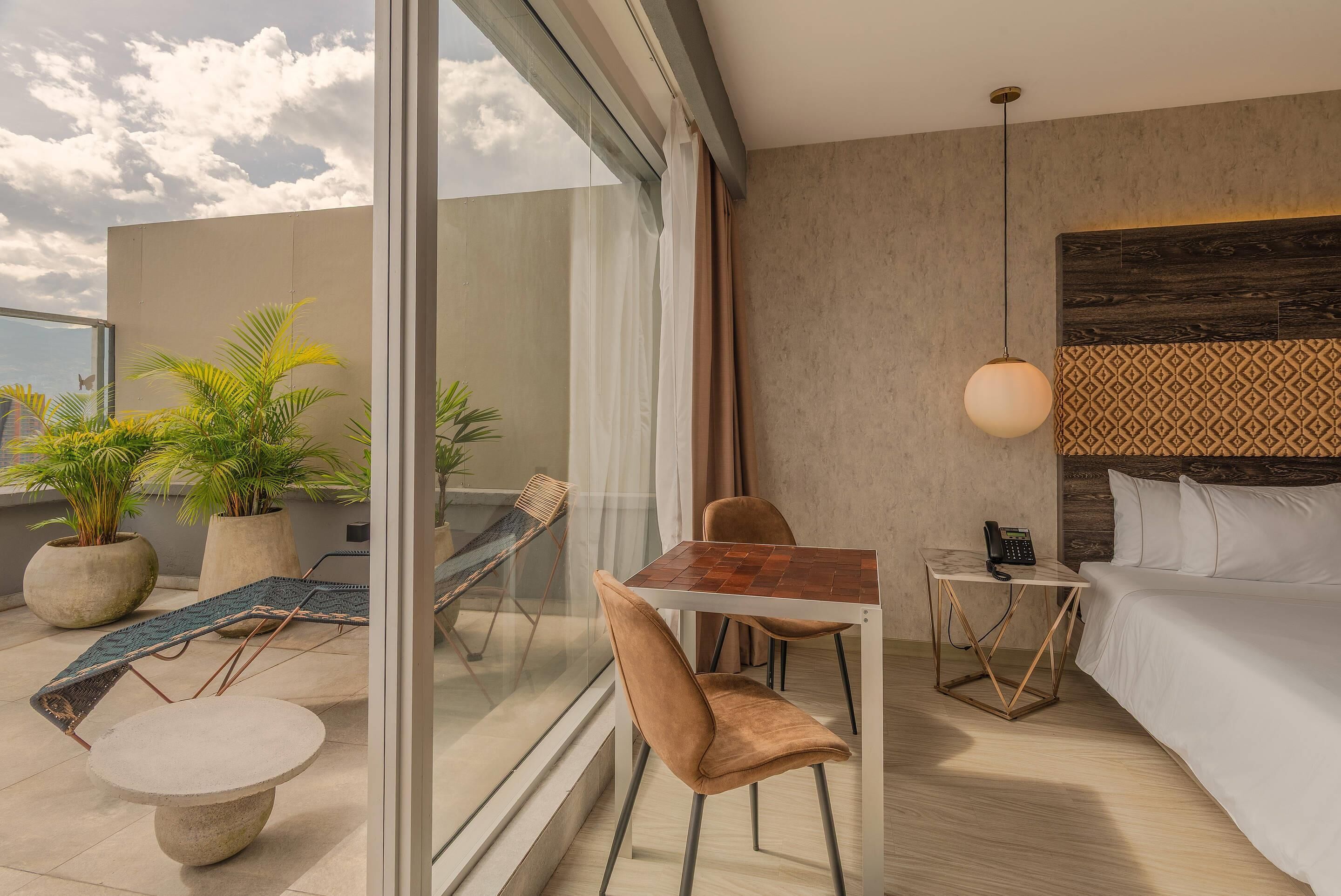 Quarto de hotel moderno com parede de vidro, cama, mesa de cabeceira com luminária, pequena mesa com cadeiras e plantas em vasos.