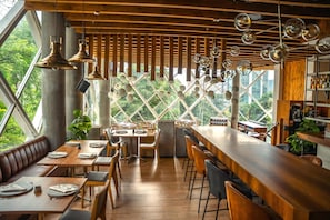 A restaurant with wooden tables and benches, a bar area, and large windows offering a view of greenery. - V Grand Hotel, a member of Radisson Individuals (Medellín)