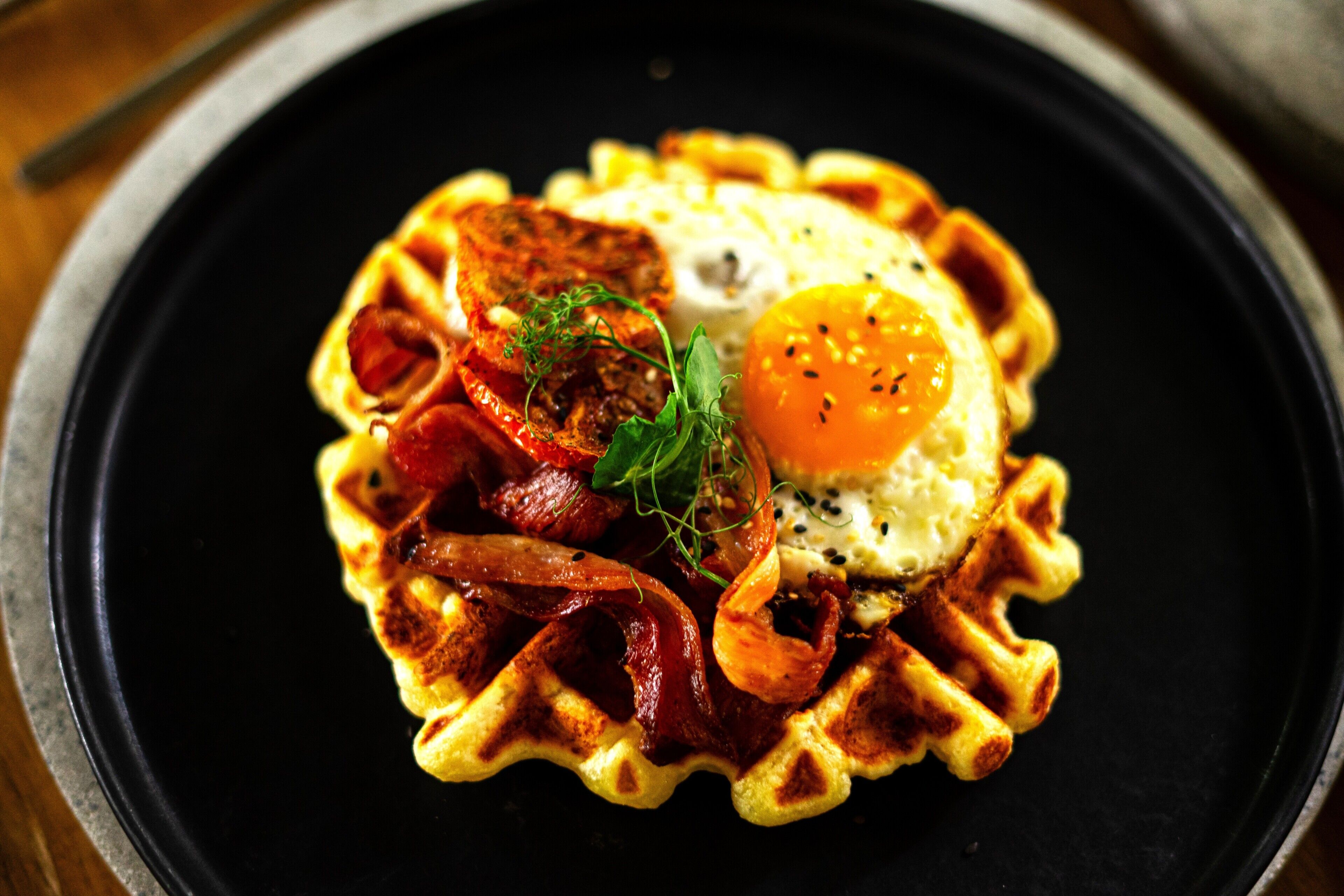 A waffle with a fried egg and bacon on a black plate.