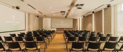 A conference room with rows of chairs facing a projection screen, a large screen displaying the hotel name, and a modern ceiling with recessed lighting.