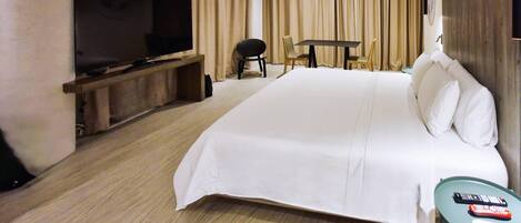 A large bed with white linens, a flat-screen TV on a wooden stand, a small table with a chair, and a remote control on the table.
