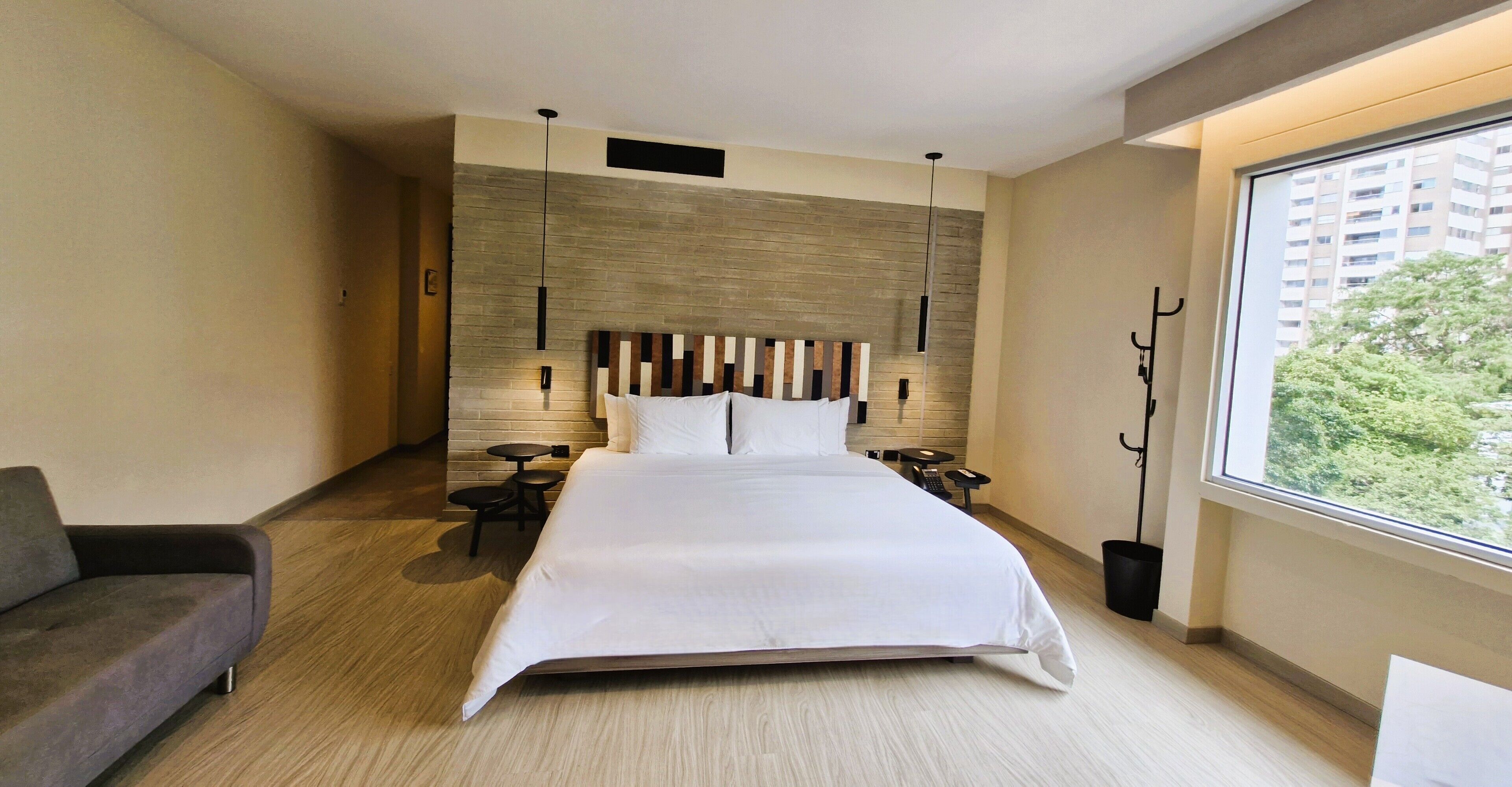 Una habitación de hotel moderna con una cama grande, un sofá, una mesita y un ventanal con vistas a edificios y árboles.
