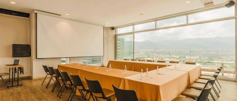 A conference room with a long table, chairs, a projector screen, and a view of a cityscape through large windows.