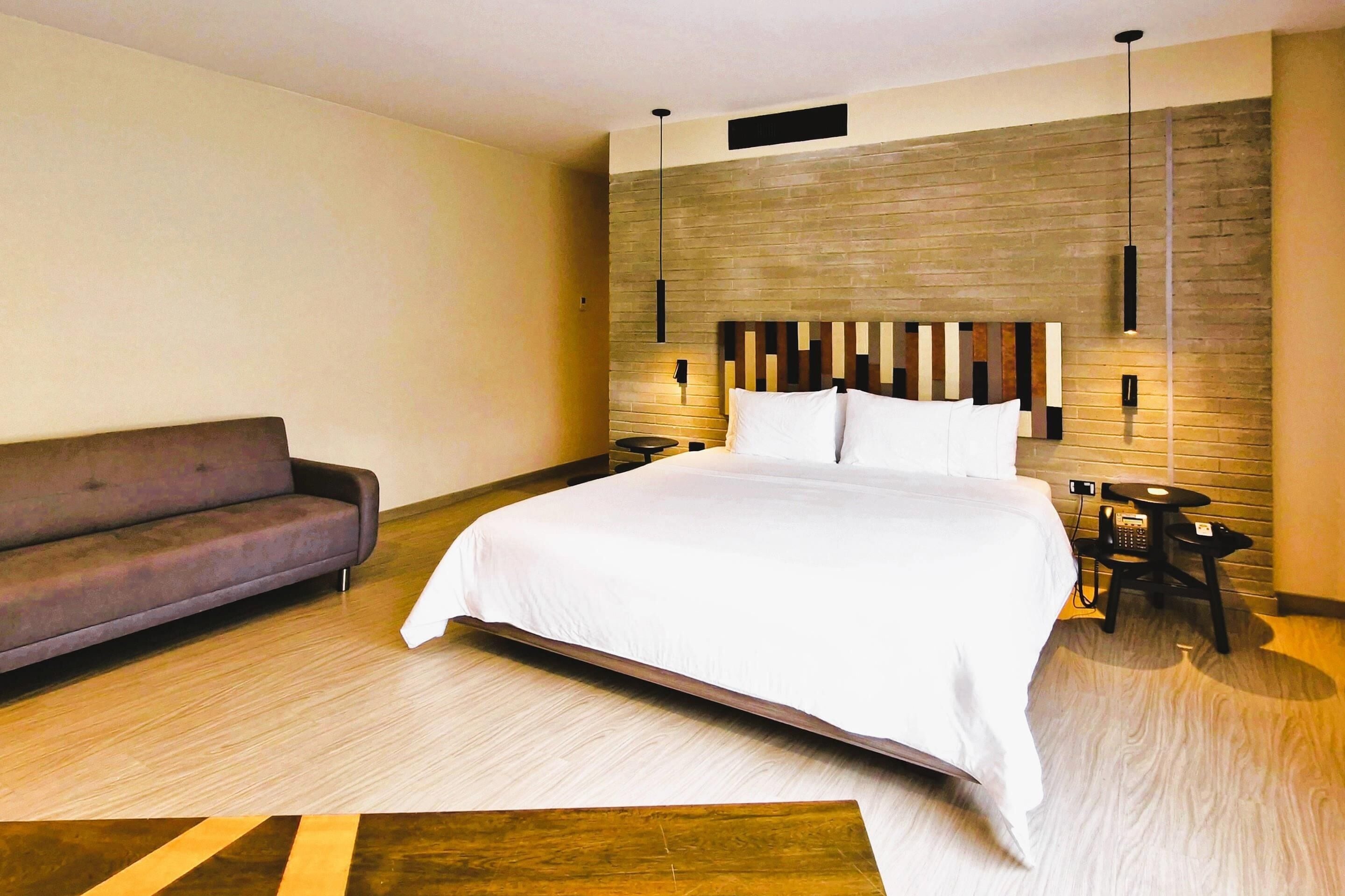 A modern hotel room with a large bed, a sofa, a wooden accent wall, and a small table with a lamp.