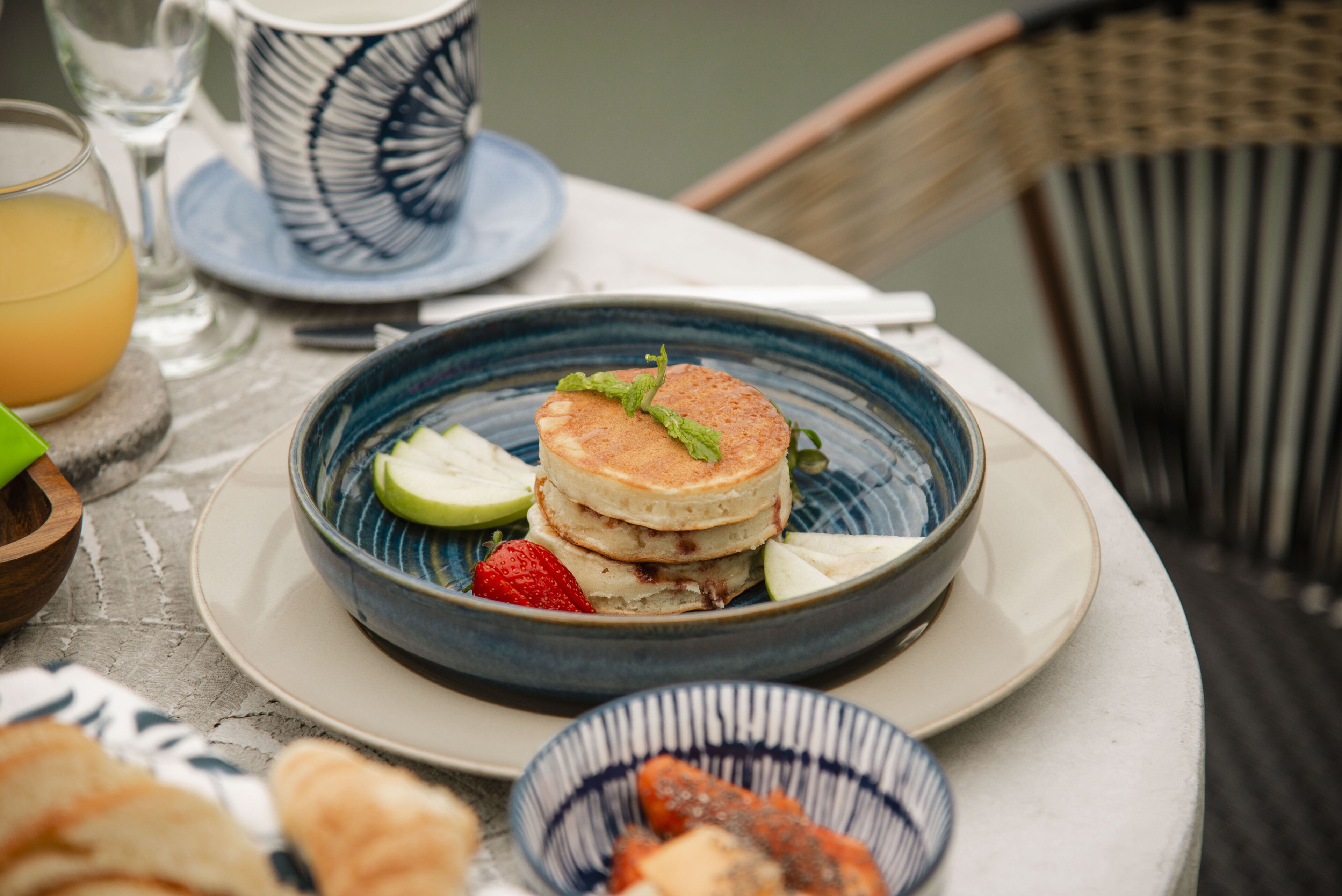 A breakfast setting with pancakes on a blue plate, accompanied by a glass of orange juice, a cup, and a bowl of fruit.