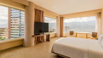 Habitación de hotel con una cama grande, televisión, escritorio con sillas y vistas a la ciudad.