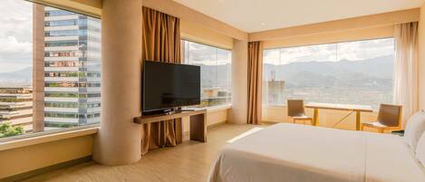 Habitación de hotel con una cama grande, televisión, escritorio con sillas y vistas a la ciudad.