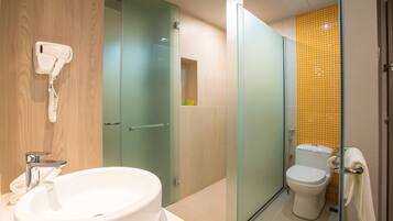 A bathroom with a glass-enclosed shower, a toilet with a yellow-tiled wall, and a sink with a hairdryer mounted on the wall.