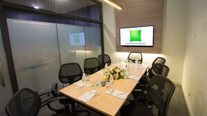 Un salón de conferencias con una larga mesa, sillas, un televisor mostrando 'ibis styles Kuala Lumpur Sri Damansara' y una decoración floral central.