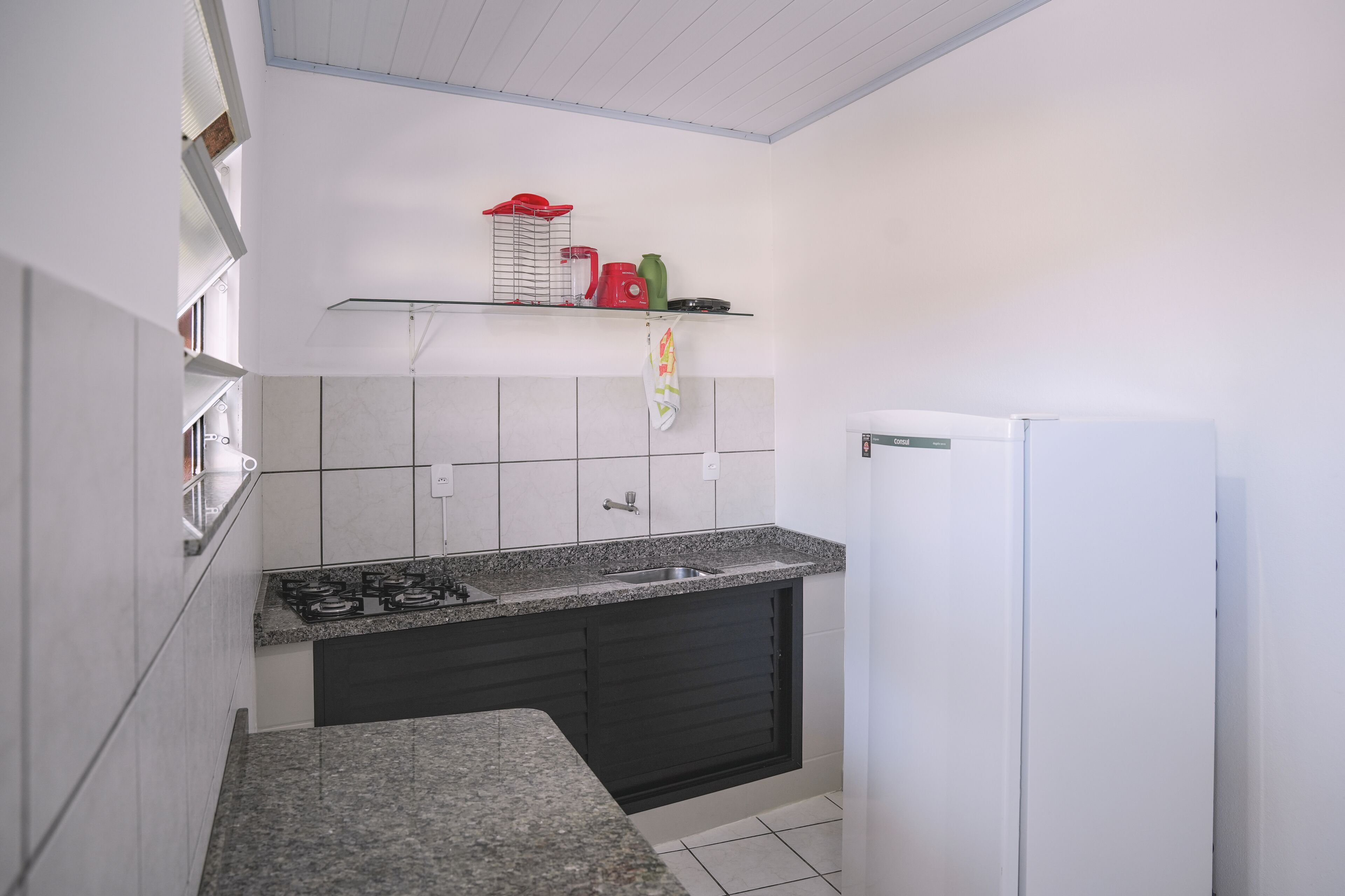 Uma cozinha compacta com bancada de granito, fogão embutido, geladeira branca e uma pequena prateleira com utensílios de cozinha.