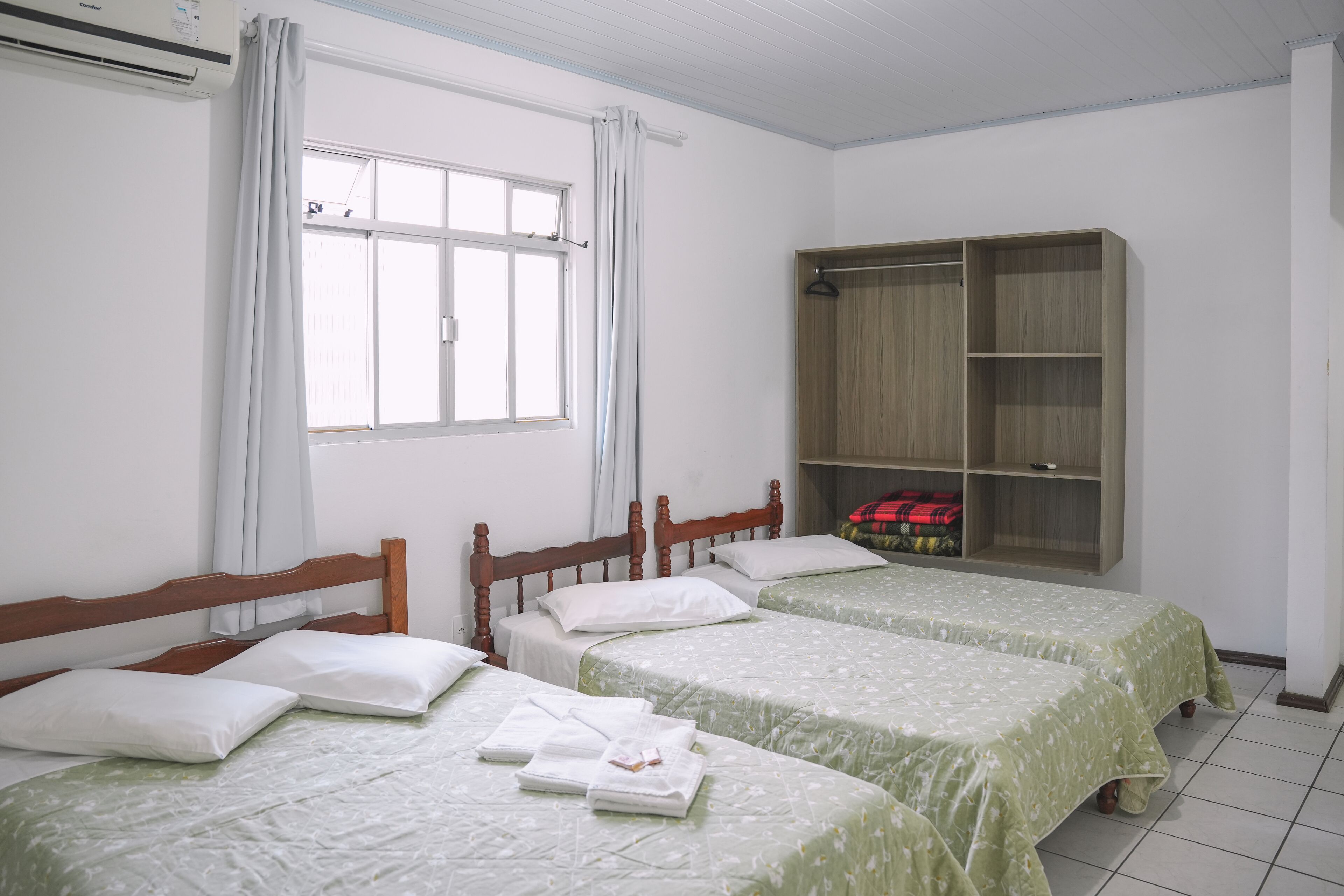 A room with two single beds, a wooden wardrobe, a window with curtains, and a tiled floor.