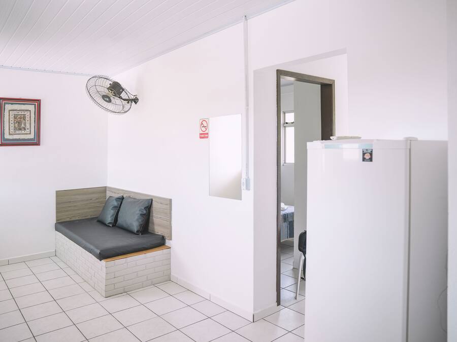 Una habitación pequeña y limpia con una cama, un ventilador de pared, un refrigerador y una puerta que da a otra habitación.