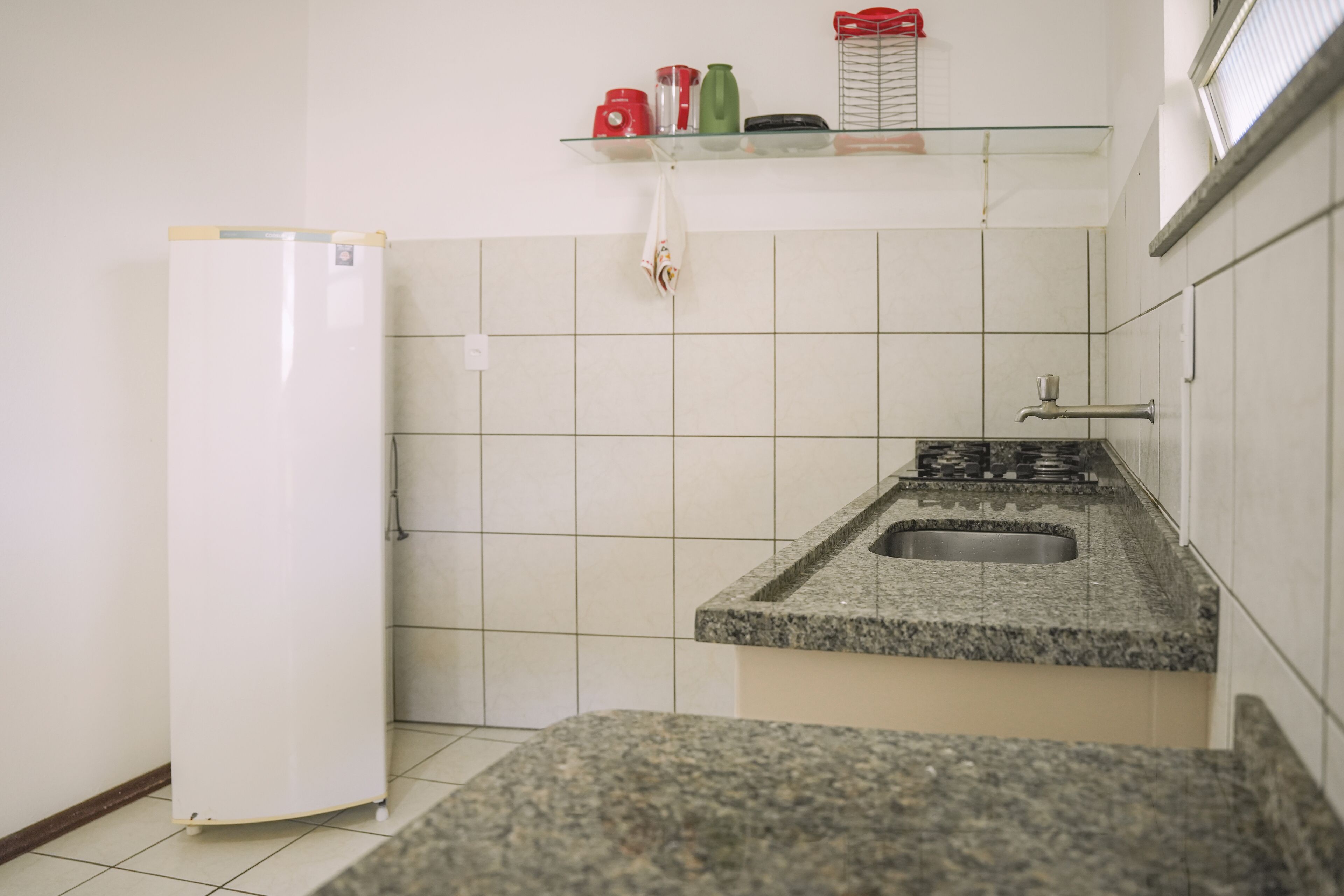A small kitchen with white tiles, a granite countertop, a stainless steel sink, and a white refrigerator.