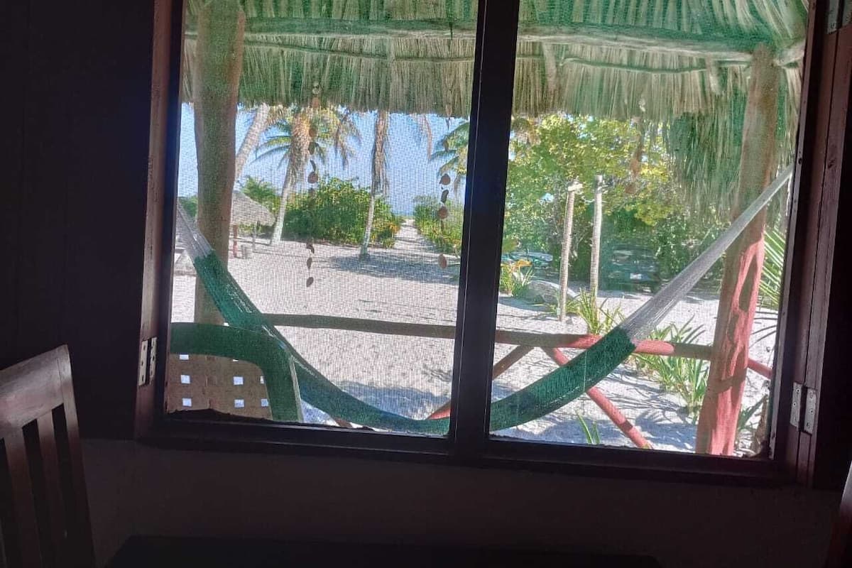 A hammock tied between two wooden posts, a wooden chair, and a view of a beach with palm trees and a clear sky.