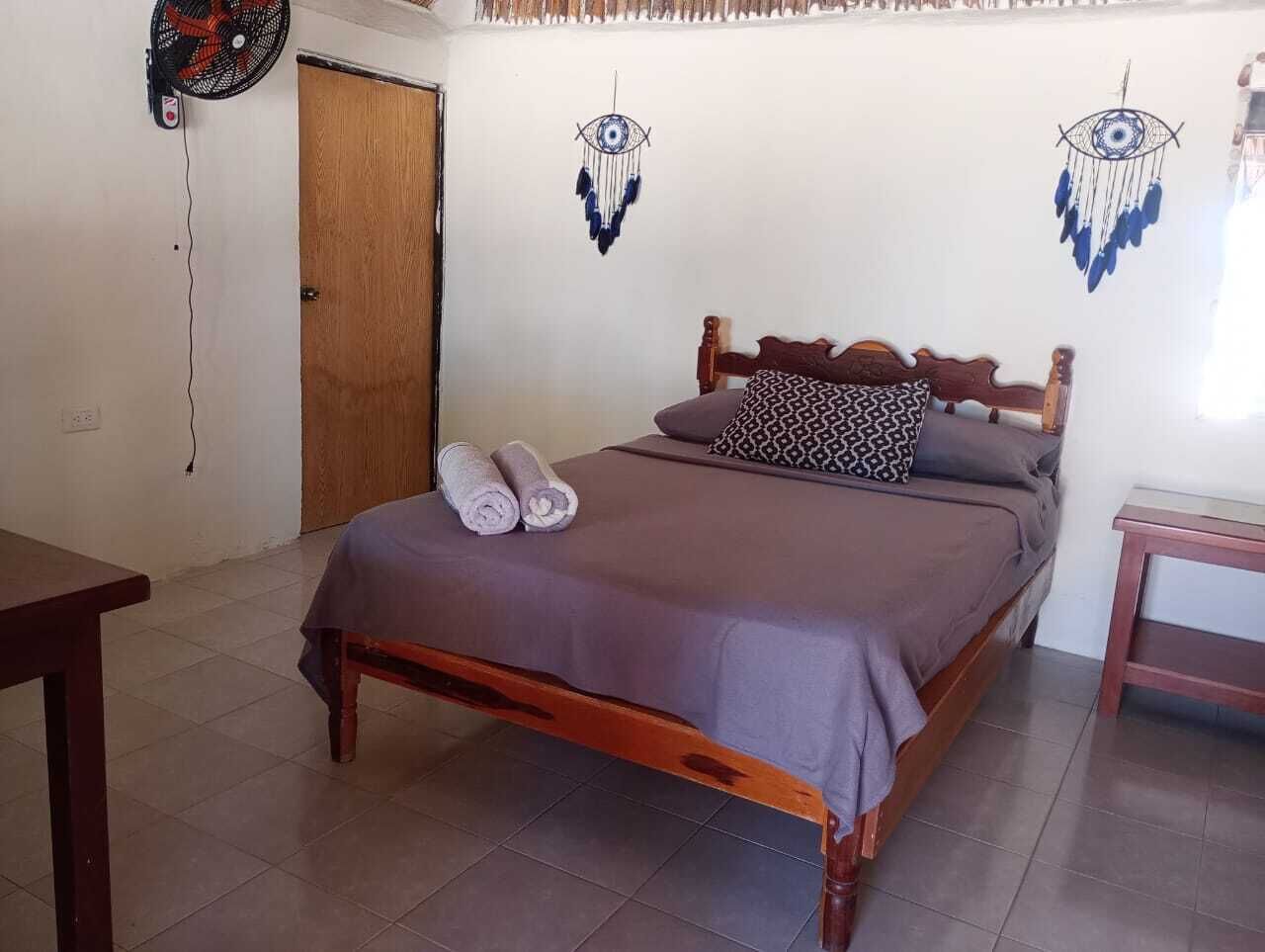 Una cama individual con estructura de madera, una colcha gris y dos toallas enrolladas. Se observan dos adornos en la pared que se asemejan a ojos, una puerta de madera y una pequeña mesa con una silla.